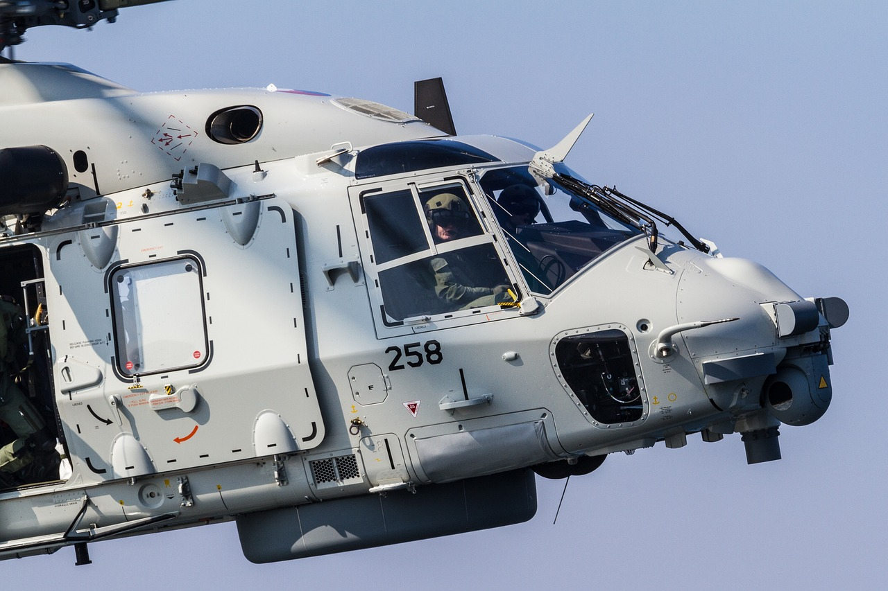 Premier plan d'un hélicoptère NH90 gris en vol, montrant le cockpit avec un pilote visible à l'intérieur.