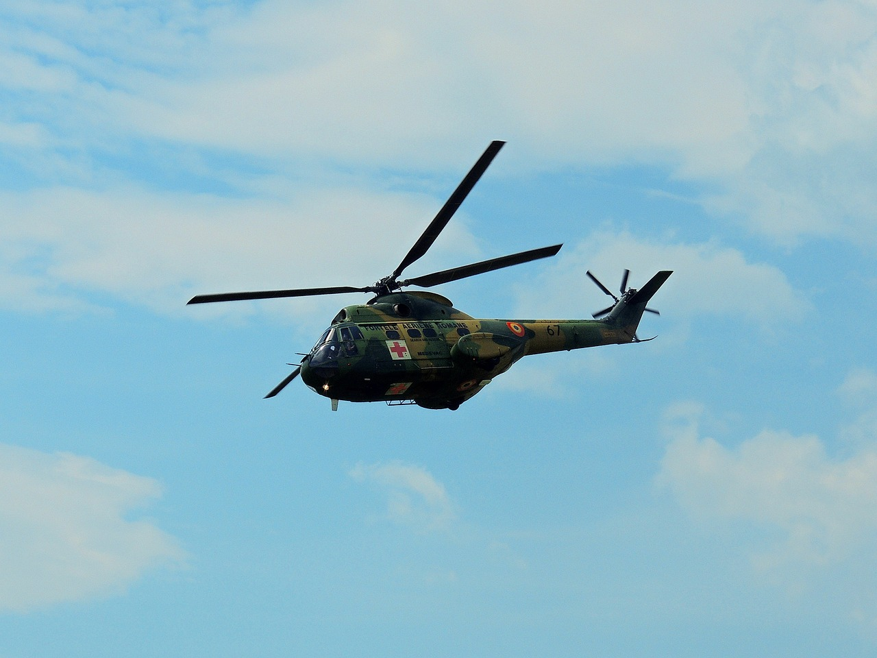 Hélicoptère militaire Puma camouflé en vol, arborant une croix rouge sur fond blanc, avec un ciel partiellement nuageux en arrière-plan.