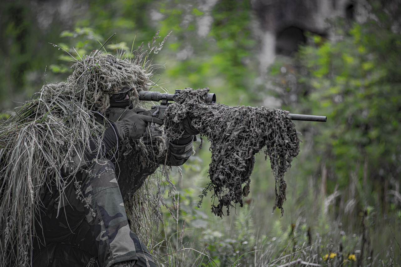 Un soldat en tenue de camouflage ghillie vise avec précision à travers son fusil équipé d'une lunette, caché dans la végétation dense d'une forêt, prêt pour une mission de sniper.