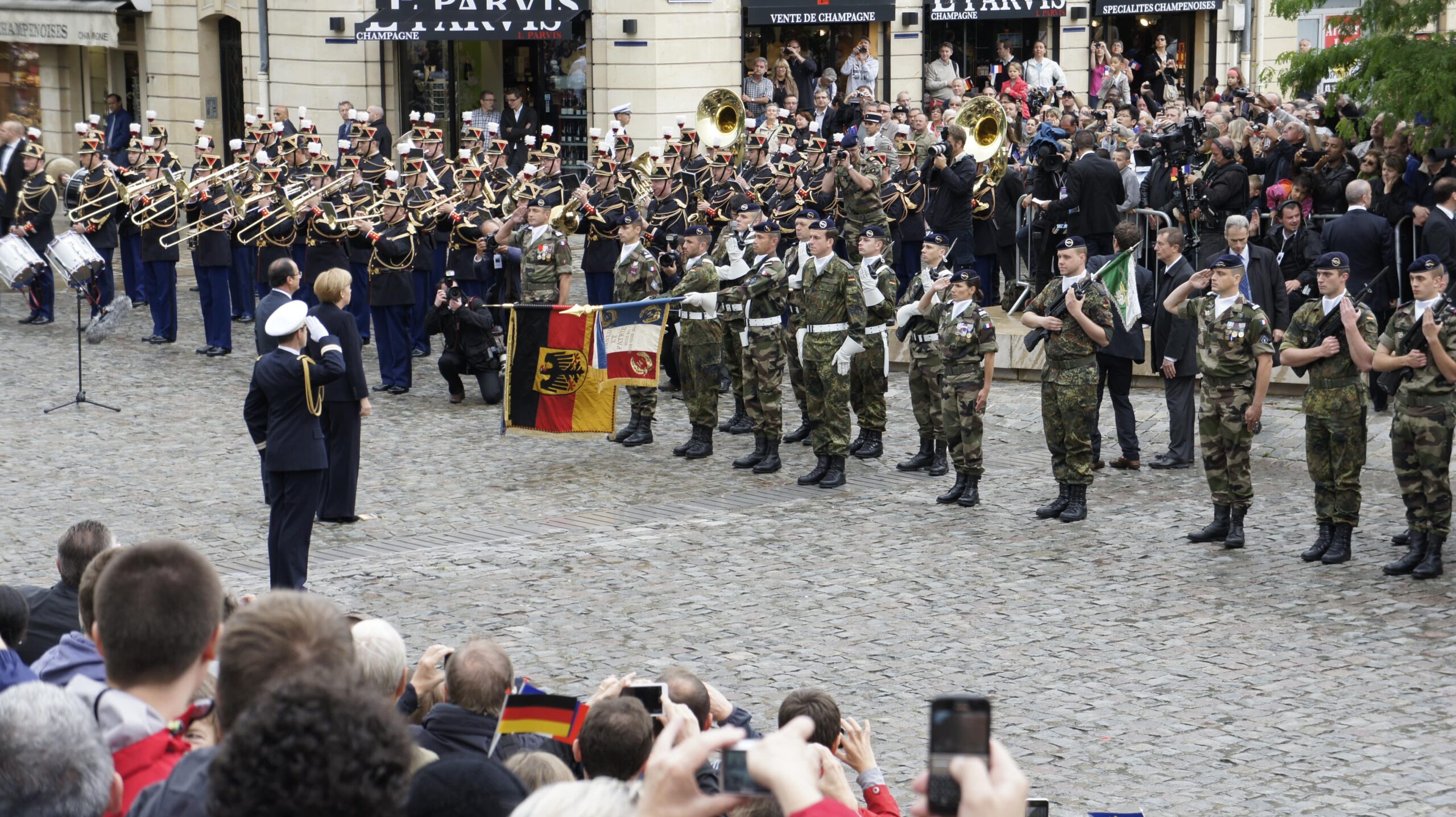 La Brigade Franco-Allemande (BFA) défile en tenue d'apparat devant une foule nombreuse, avec des drapeaux français et allemand. Une fanfare militaire joue en arrière-plan. Des dignitaires assistent à la cérémonie sur une place pavée, symbolisant l'amitié franco-allemande.