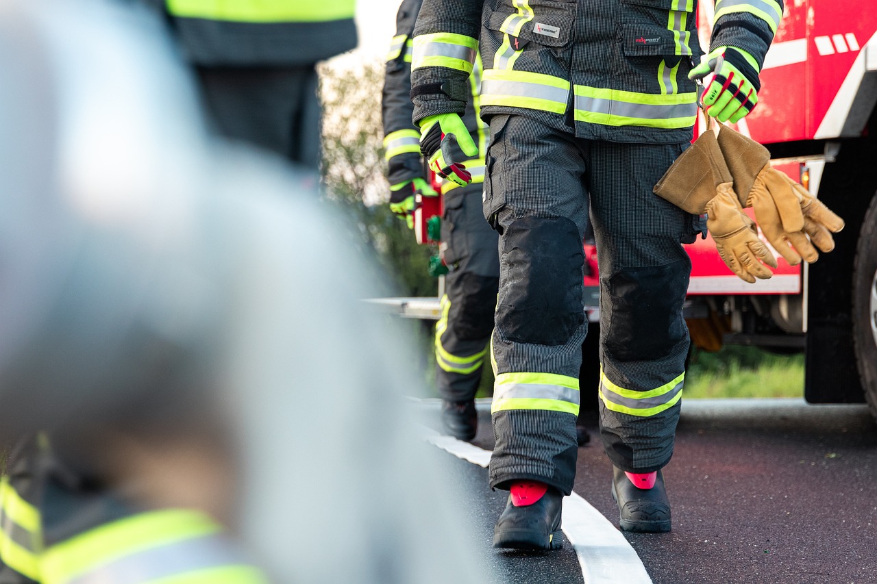 Des pompiers en tenue de protection, avec des bandes réfléchissantes, marchent vers un camion de pompiers rouge. L'un d'eux tient une paire de gants en cuir. La photo se concentre sur le bas de leur corps, mettant en avant leur équipement de sécurité.