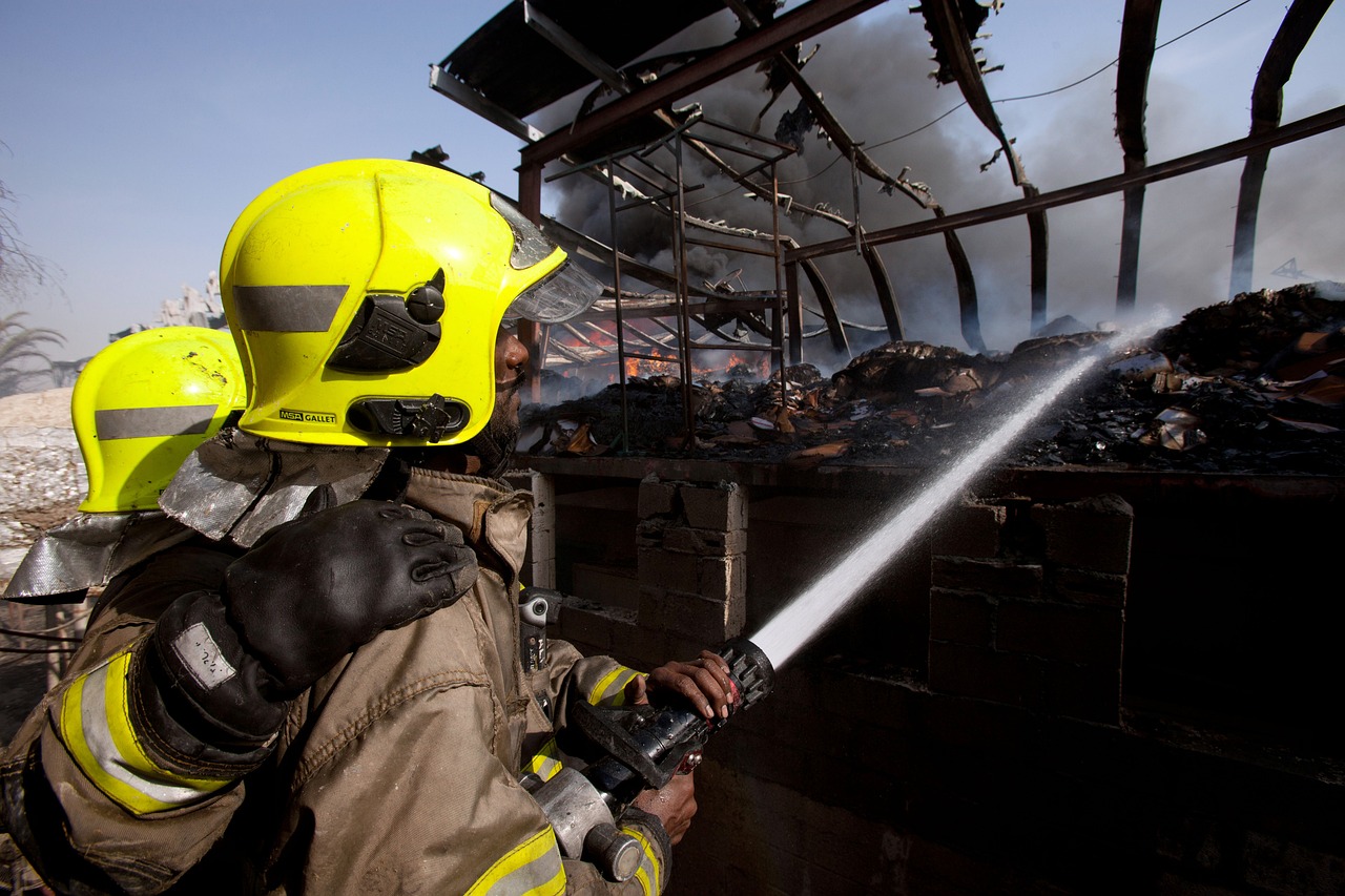 Des pompiers, équipés de casques jaunes et de combinaisons ignifuges, combattent un incendie dans un bâtiment en flammes. L'un d'eux tient une lance à incendie, projetant de l'eau sur les débris en feu, tandis que l'autre le soutient. La structure du bâtiment est partiellement effondrée, et des fumées épaisses s'élèvent.