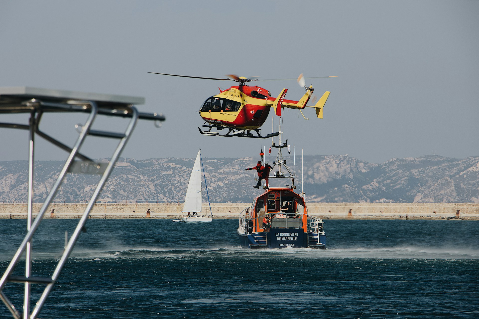 Un hélicoptère de secours jaune et rouge effectue une opération de sauvetage au-dessus d'un bateau de sauvetage nommé "La Bonne Mère de Marseille". Un secouriste est en train de descendre en rappel depuis l'hélicoptère vers le bateau. En arrière-plan, on aperçoit des montagnes et un voilier naviguant sur l'eau. La scène se déroule en mer, illustrant une mission de sauvetage maritime.