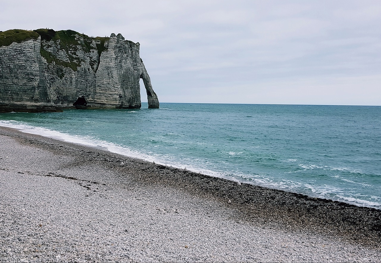 Plage de galets en Normandie avec vue sur une falaise rocheuse et une arche naturelle se jetant dans la mer turquoise, sous un ciel nuageux.