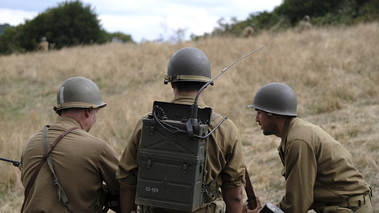 Trois soldats en uniforme militaire se tiennent dos à la caméra, le soldat du centre porte un gros équipement radio