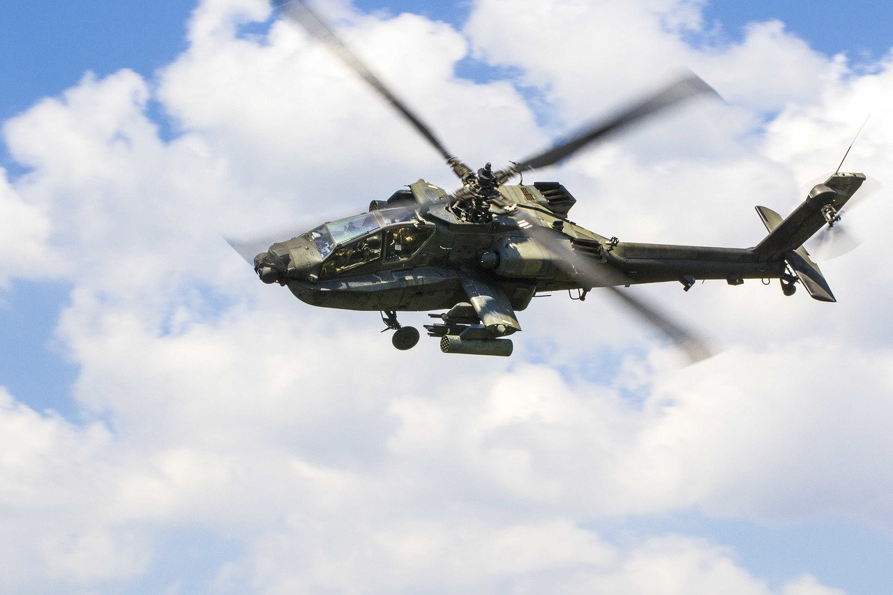 Hélicoptère militaire Apache en vol, avec ses pales en mouvement et un ciel nuageux en arrière-plan.
