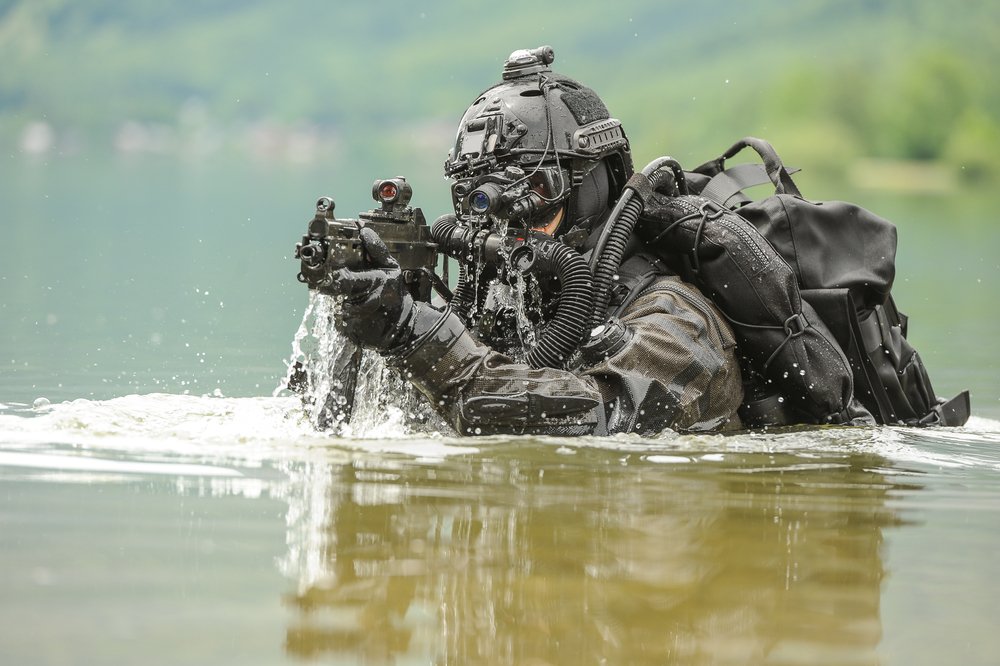 Un soldat en tenue tactique émerge de l'eau, tenant une arme, avec une forêt en arrière-plan.