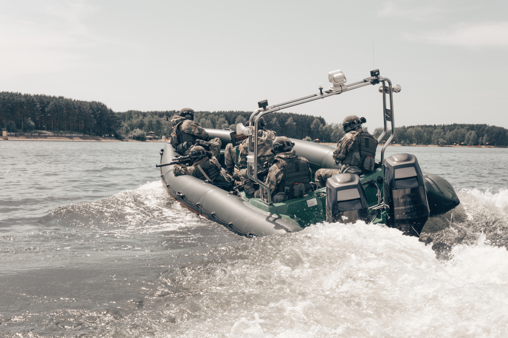 Des soldats en uniforme militaire naviguent rapidement sur un bateau pneumatique motorisé, en mission ou en exercice, avec une forêt en arrière-plan et un ciel dégagé.