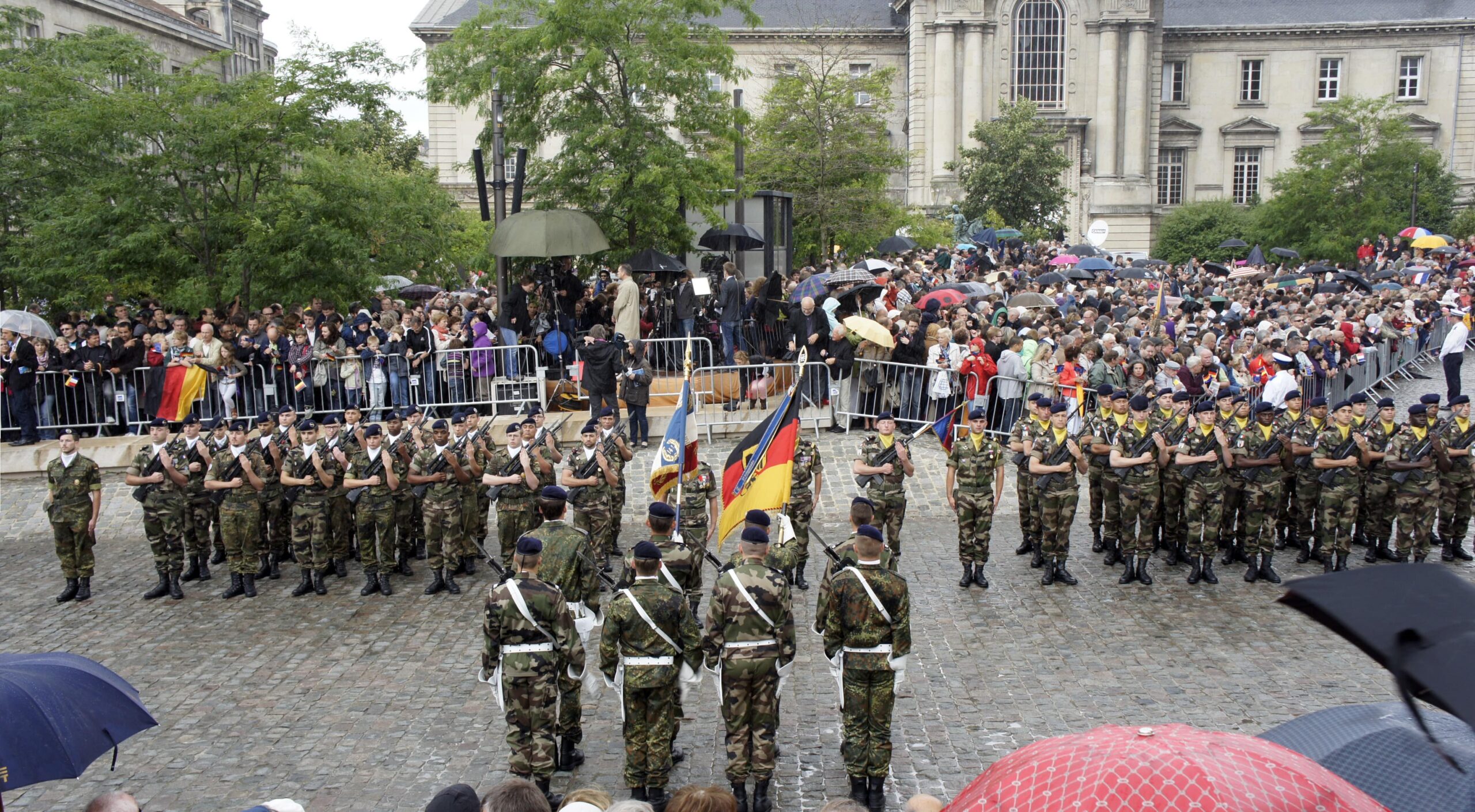 La Brigade Franco-Allemande (BFA) défile en tenue militaire devant une foule rassemblée, arborant les drapeaux français et allemand. La scène se déroule sur une place pavée, sous un ciel pluvieux, entourée de bâtiments historiques.