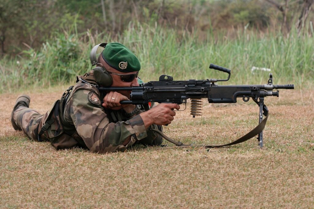 Un soldat en uniforme de camouflage est allongé sur le sol, tenant une mitrailleuse. Il porte un béret vert, des lunettes de protection et un casque antibruit.