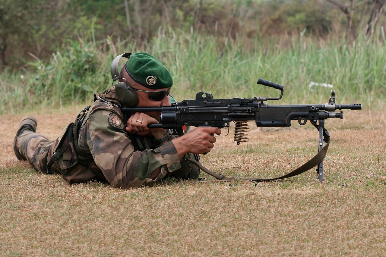 Un soldat en uniforme de camouflage est allongé sur le sol, tenant une mitrailleuse. Il porte un béret vert, des lunettes de protection et un casque antibruit.