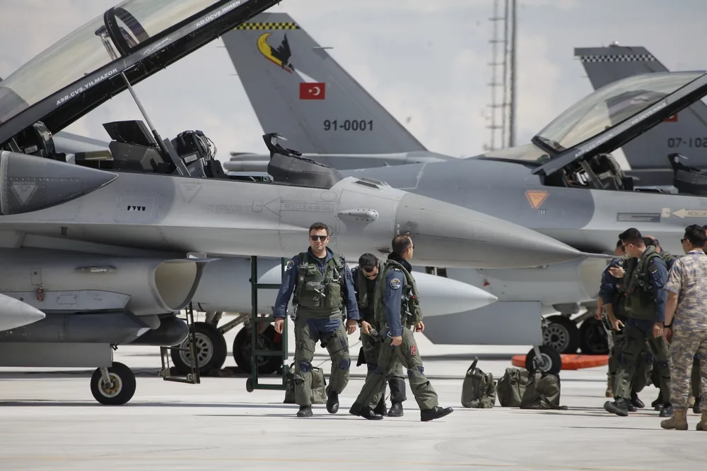 Pilotes militaires en tenue de vol effectuant un briefing devant des avions de chasse sur un tarmac.