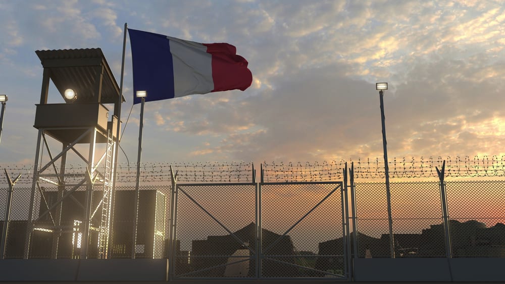 Drapeau français flottant au-dessus de l'entrée d'une caserne militaire avec mirador et clôture barbelée.