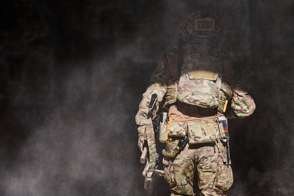 Militaire équipé d'un gilet tactique et d'un fusil d'assaut, vu de dos dans la poussière lors d'un entraînement de terrain.