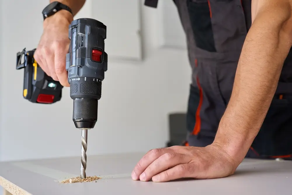 Mains d'un opérateur en tenue d'atelier perçant verticalement une planche de bois avec une perceuse-visseuse sans fil